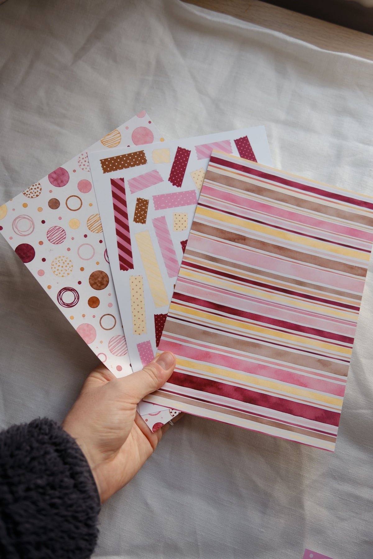 A hand holding three sheets of decorative washi tape with different patterns, including vertical stripes and polka dots, over a white fabric background.